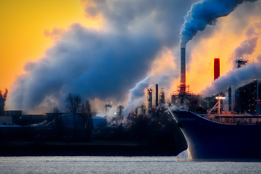 Cargo ship passing dockland factories, all visibly emitting exhaust fumes.