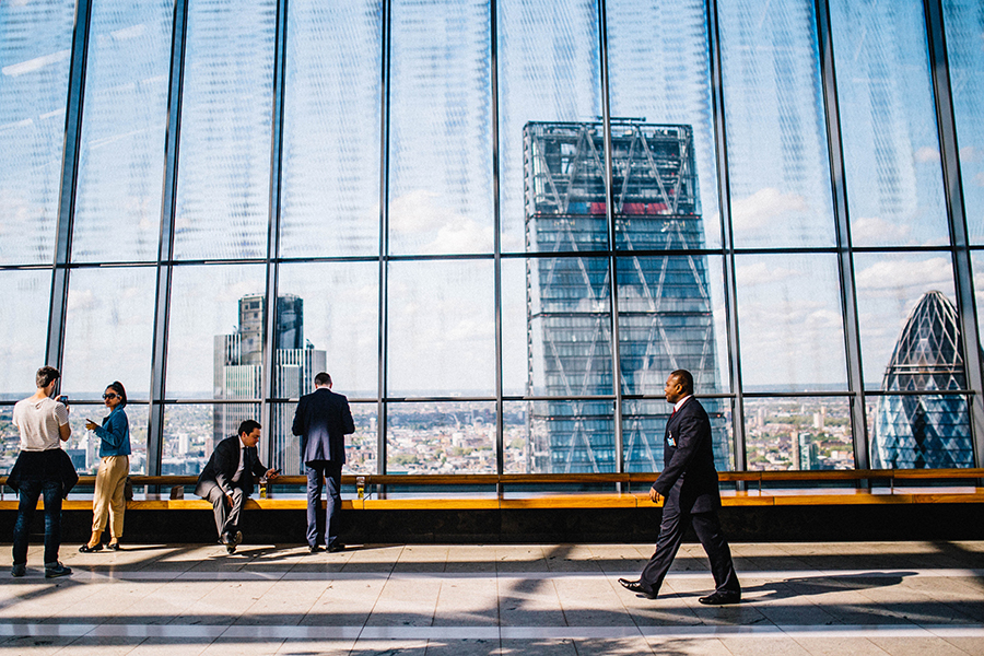 View of a floor of a skyscraper with office workers moving around and a city skyline appearing in the glass wall behind them.