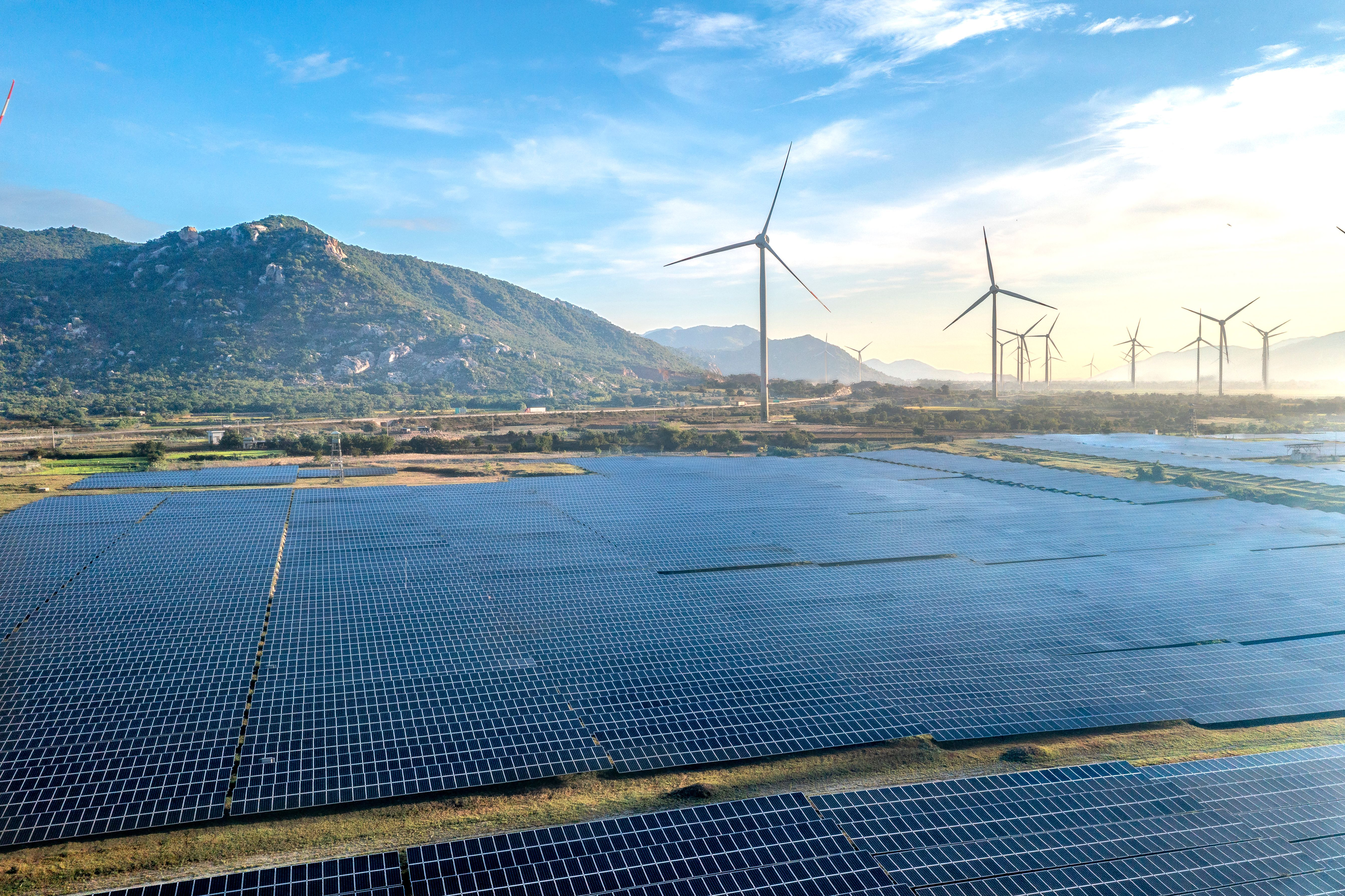 Fields of solar panels and wind turbines under a blue sky.